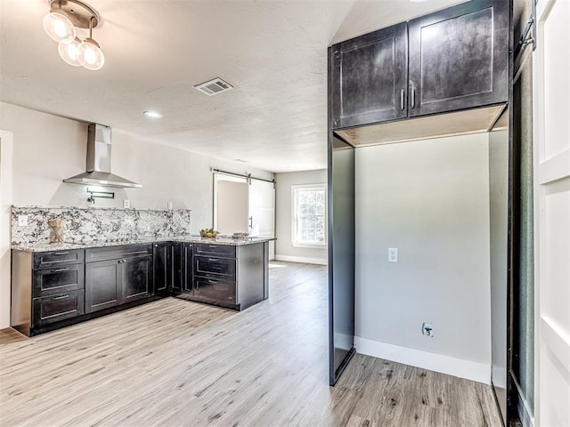 kitchen featuring wall chimney range hood, a barn door, light hardwood / wood-style floors, light stone counters, and kitchen peninsula