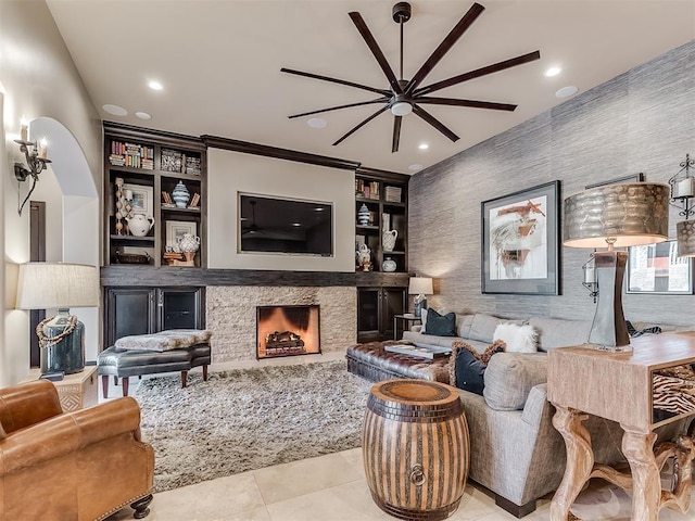 living room with built in shelves, ceiling fan, crown molding, light tile patterned floors, and a stone fireplace