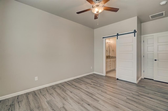 unfurnished bedroom with ceiling fan, a barn door, light wood-type flooring, and connected bathroom