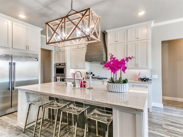 kitchen featuring wall chimney range hood, a notable chandelier, appliances with stainless steel finishes, white cabinetry, and light wood-type flooring