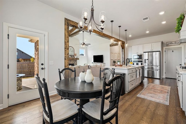 dining area featuring ceiling fan with notable chandelier, dark wood-type flooring, sink, vaulted ceiling with beams, and a stone fireplace