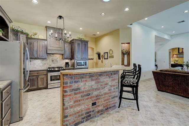 kitchen with an inviting chandelier, decorative backsplash, an island with sink, dark brown cabinetry, and stainless steel appliances