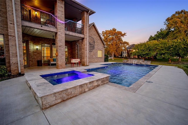 pool at dusk featuring pool water feature, a patio area, and an in ground hot tub