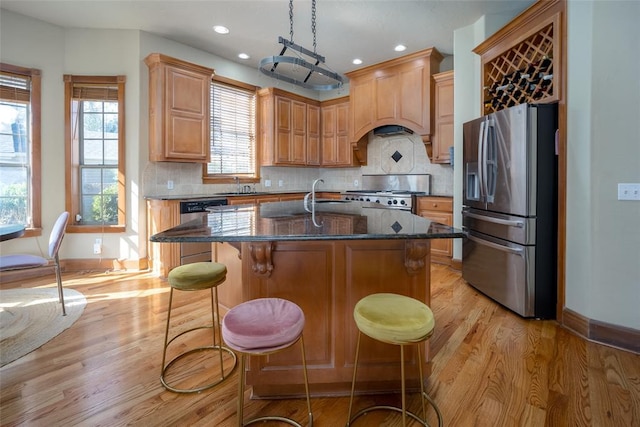 kitchen with stainless steel appliances, dark stone countertops, an island with sink, premium range hood, and a breakfast bar area