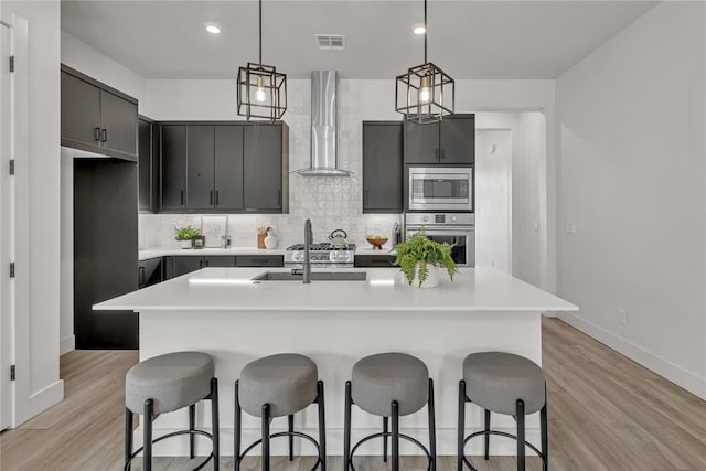 kitchen with wall chimney exhaust hood, light hardwood / wood-style floors, an island with sink, and appliances with stainless steel finishes