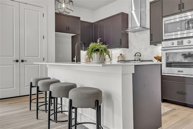 kitchen featuring appliances with stainless steel finishes, light hardwood / wood-style flooring, a kitchen island with sink, and wall chimney range hood