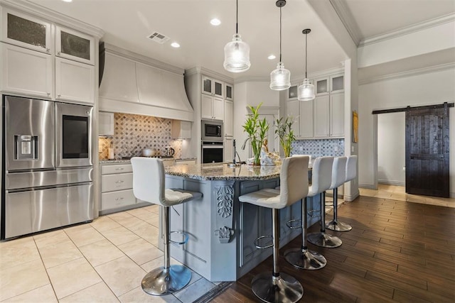 kitchen featuring premium range hood, a center island with sink, white cabinets, a barn door, and appliances with stainless steel finishes