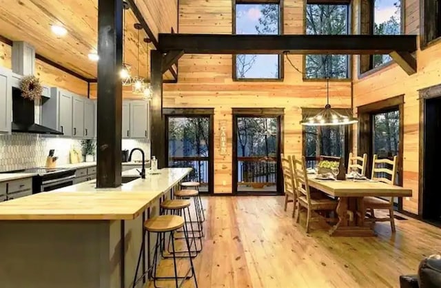 kitchen with wall chimney exhaust hood, decorative light fixtures, stainless steel electric stove, wooden walls, and light wood-type flooring