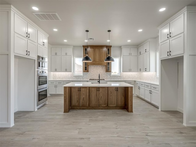 kitchen with hanging light fixtures, an island with sink, tasteful backsplash, light hardwood / wood-style floors, and white cabinetry