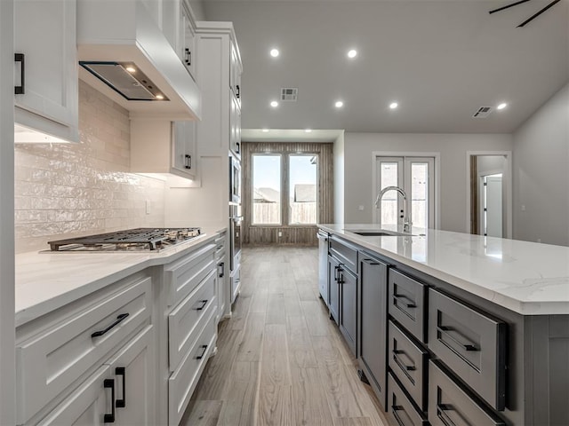 kitchen with white cabinetry, sink, a kitchen island with sink, light stone counters, and custom range hood