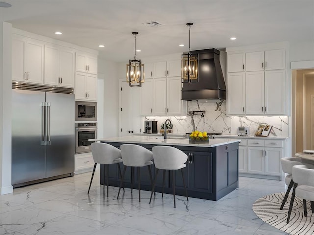 kitchen featuring premium range hood, pendant lighting, white cabinetry, a kitchen island with sink, and built in appliances