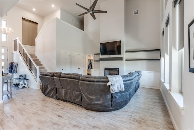 living room featuring light wood-type flooring, a towering ceiling, a brick fireplace, and ceiling fan