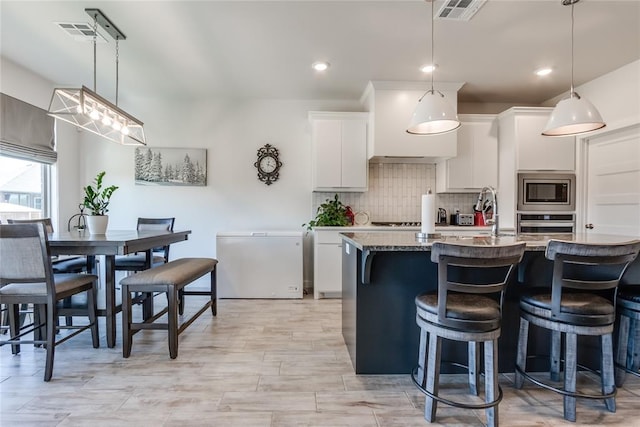 kitchen with hanging light fixtures, stainless steel appliances, white cabinetry, and a kitchen island with sink