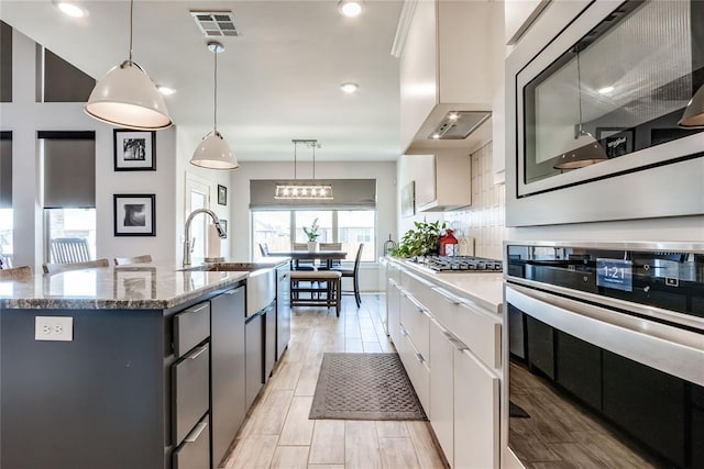 kitchen with white cabinets, decorative backsplash, an island with sink, appliances with stainless steel finishes, and decorative light fixtures