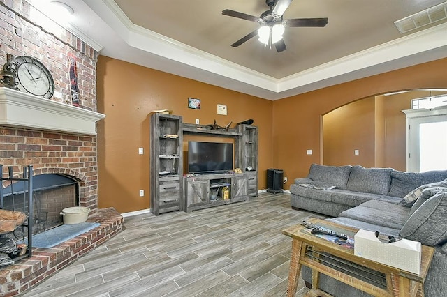 living room featuring a raised ceiling, a brick fireplace, ceiling fan, light wood-type flooring, and ornamental molding