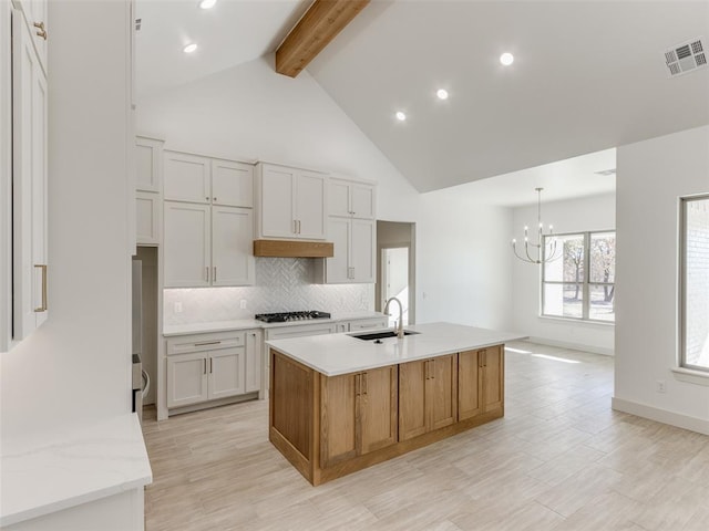 kitchen with white cabinetry, sink, beamed ceiling, stainless steel gas stovetop, and a kitchen island with sink