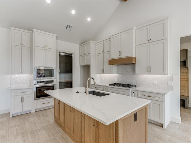 kitchen with appliances with stainless steel finishes, vaulted ceiling, a kitchen island with sink, sink, and white cabinets