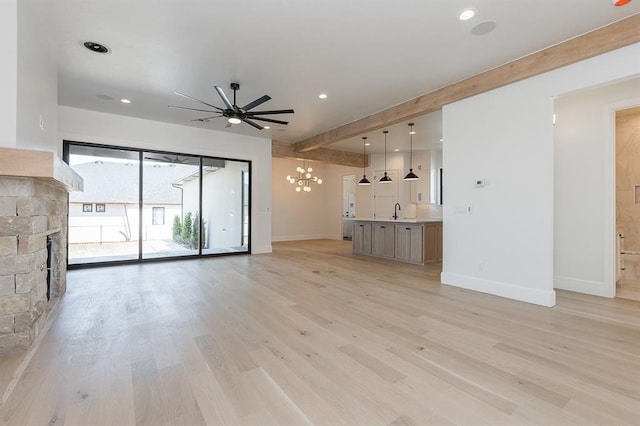 unfurnished living room featuring sink, a stone fireplace, light hardwood / wood-style flooring, beamed ceiling, and ceiling fan with notable chandelier