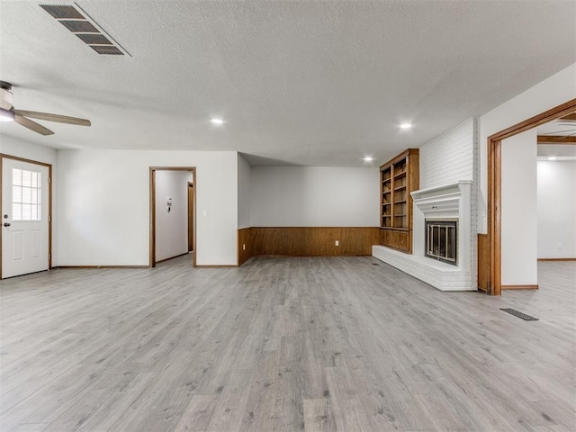 unfurnished living room with built in shelves, ceiling fan, a brick fireplace, light hardwood / wood-style flooring, and a textured ceiling