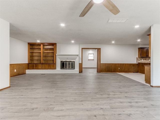 unfurnished living room featuring light wood-type flooring, a brick fireplace, ceiling fan, wooden walls, and built in features