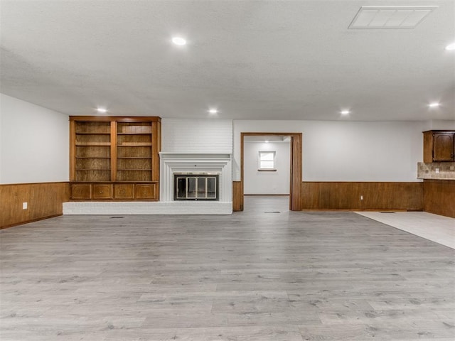 unfurnished living room featuring built in shelves, a brick fireplace, light hardwood / wood-style flooring, wood walls, and a textured ceiling