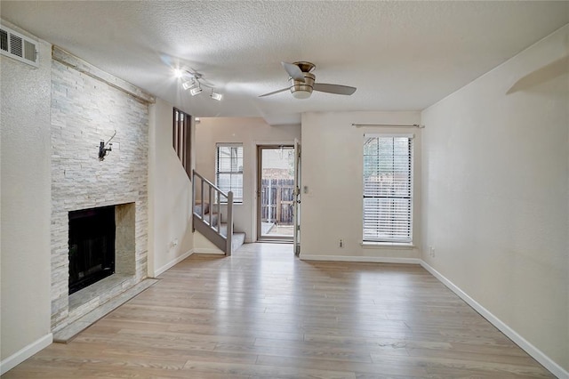 unfurnished living room with ceiling fan, a stone fireplace, a textured ceiling, and light hardwood / wood-style flooring