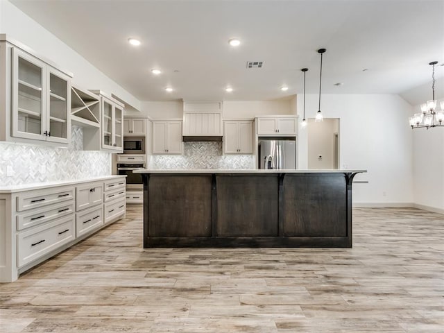 kitchen with appliances with stainless steel finishes, backsplash, a large island with sink, decorative light fixtures, and light hardwood / wood-style flooring