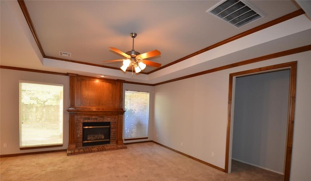 unfurnished living room featuring a raised ceiling, a brick fireplace, ceiling fan, ornamental molding, and light colored carpet