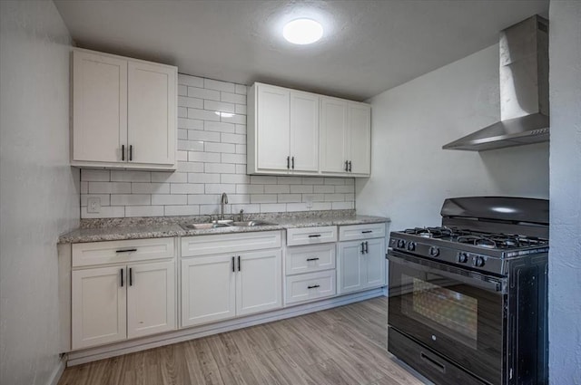 kitchen with light wood-type flooring, wall chimney exhaust hood, black range with gas stovetop, sink, and white cabinetry