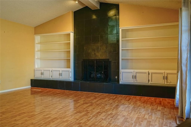 unfurnished living room featuring vaulted ceiling with beams, built in shelves, light wood-type flooring, a fireplace, and a textured ceiling