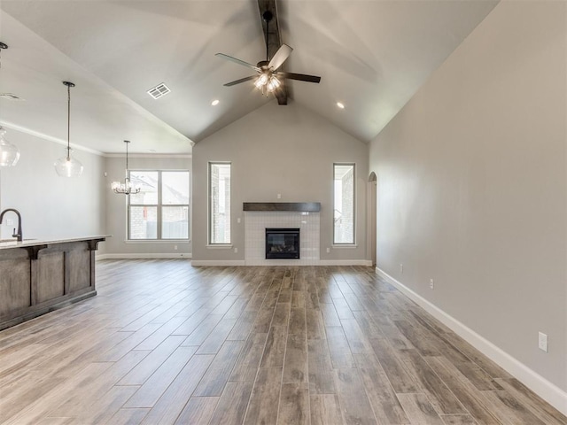 unfurnished living room with light wood-type flooring, ceiling fan with notable chandelier, sink, a tile fireplace, and vaulted ceiling with beams