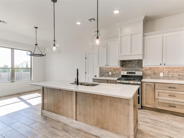 kitchen featuring stainless steel gas range oven, sink, decorative light fixtures, white cabinets, and an island with sink