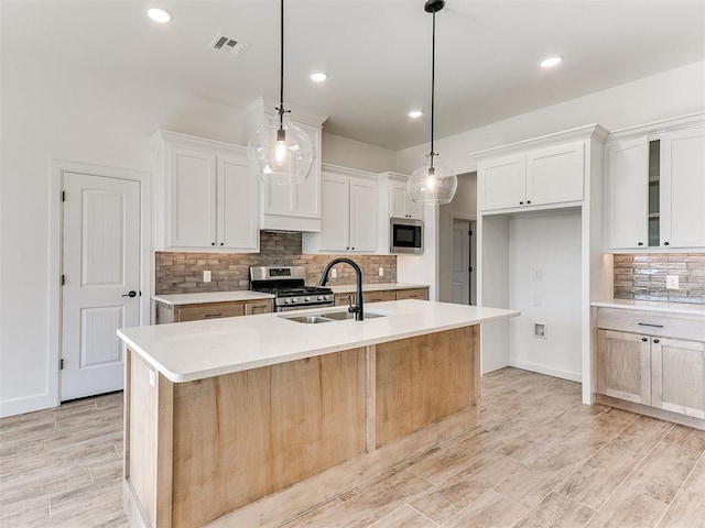 kitchen with light wood-type flooring, stainless steel appliances, a kitchen island with sink, decorative light fixtures, and white cabinets