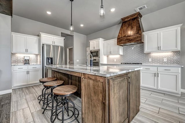 kitchen featuring custom range hood, stainless steel appliances, decorative light fixtures, a center island with sink, and white cabinetry