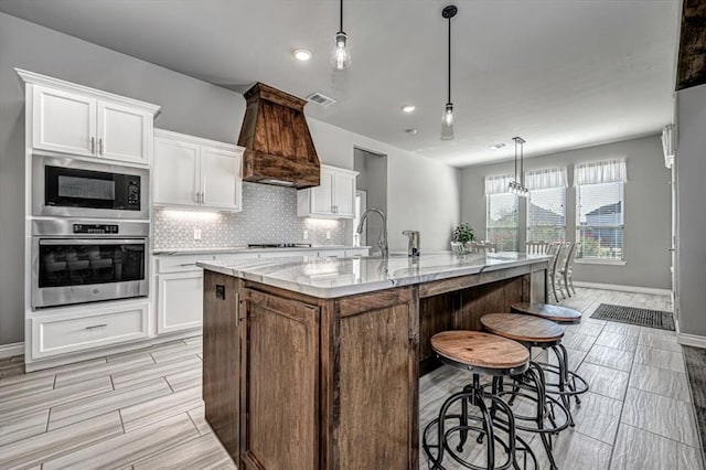 kitchen with built in microwave, white cabinetry, stainless steel oven, decorative light fixtures, and a kitchen island with sink