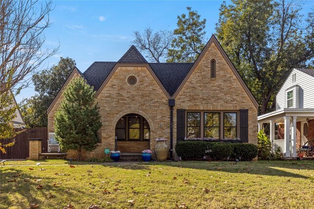 english style home with roof with shingles, brick siding, a front lawn, and fence