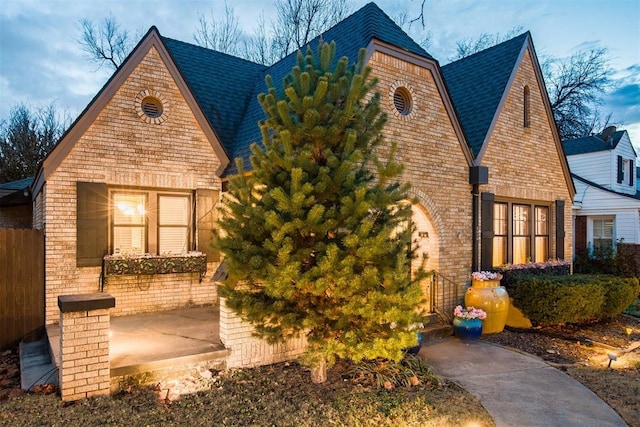 english style home featuring brick siding, roof with shingles, a patio area, and fence