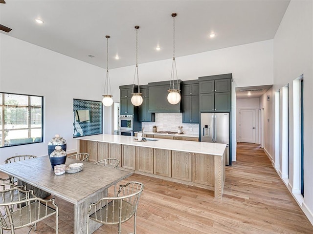 kitchen with appliances with stainless steel finishes, a towering ceiling, light hardwood / wood-style flooring, hanging light fixtures, and an island with sink