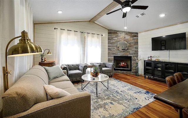 living room featuring ceiling fan, vaulted ceiling with beams, a stone fireplace, wood walls, and hardwood / wood-style flooring