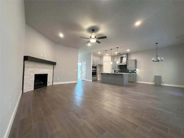 unfurnished living room featuring dark wood-type flooring, sink, a stone fireplace, and ceiling fan with notable chandelier