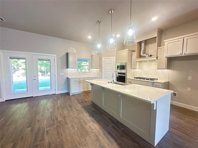 kitchen with appliances with stainless steel finishes, custom range hood, pendant lighting, dark hardwood / wood-style floors, and an island with sink