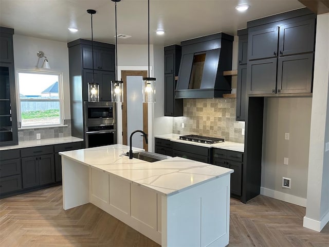 kitchen with sink, stainless steel appliances, light stone counters, premium range hood, and a kitchen island with sink