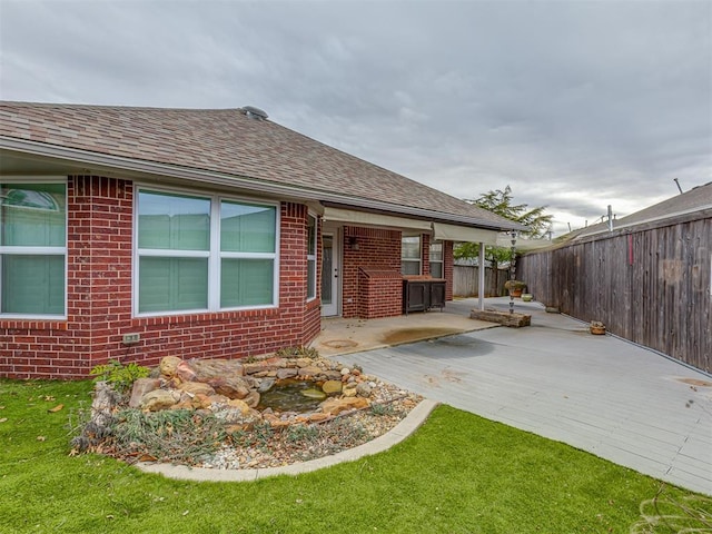 exterior space with brick siding, roof with shingles, a lawn, a patio area, and fence