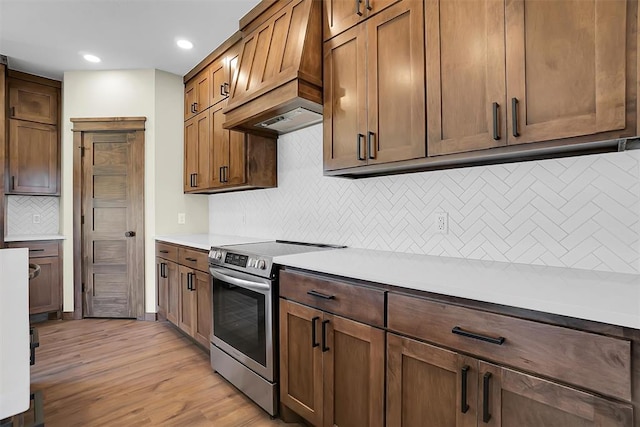 kitchen featuring backsplash, premium range hood, stainless steel electric stove, and light hardwood / wood-style floors