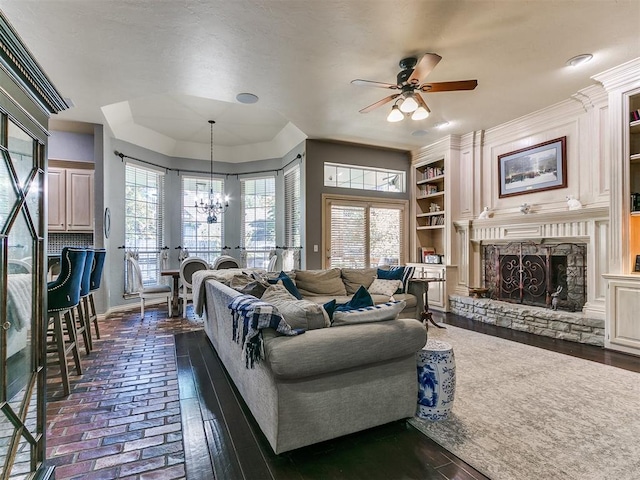 living room with ceiling fan with notable chandelier, dark hardwood / wood-style flooring, a stone fireplace, and built in shelves