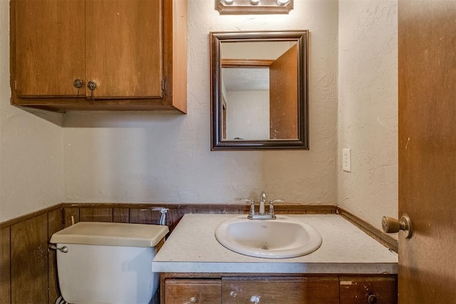 bathroom with a textured wall, toilet, vanity, and a wainscoted wall