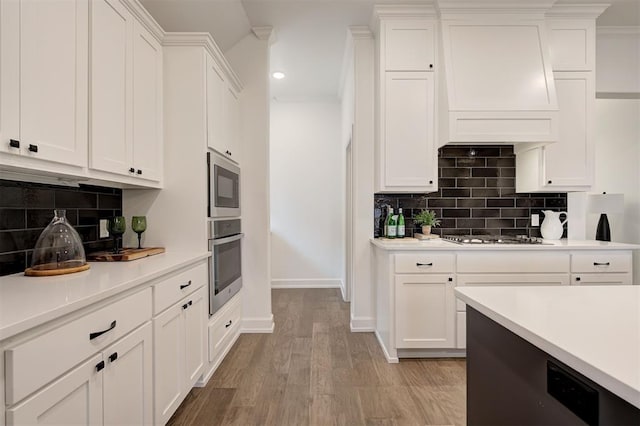 kitchen featuring custom exhaust hood, white cabinetry, light wood-type flooring, appliances with stainless steel finishes, and backsplash