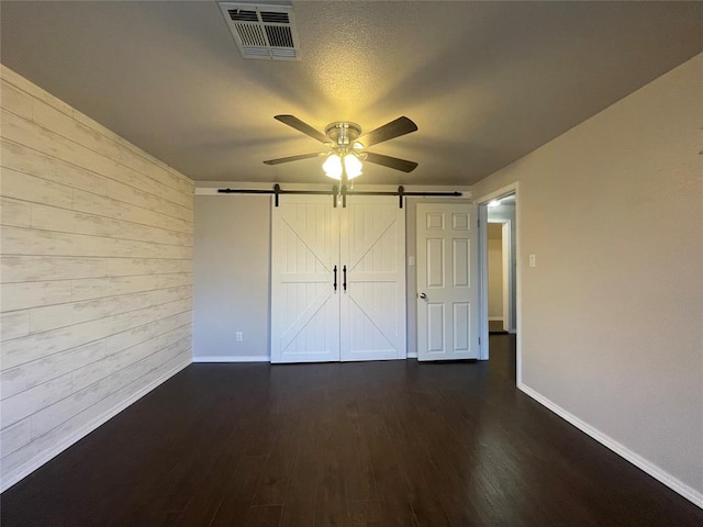 unfurnished bedroom featuring ceiling fan, a barn door, dark wood-type flooring, and wooden walls