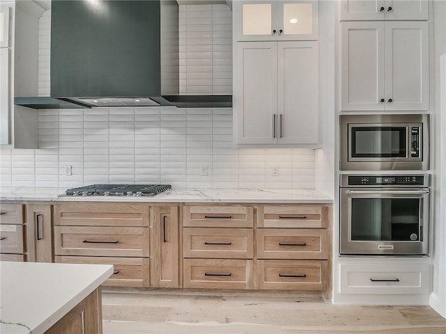 kitchen with white cabinetry, light brown cabinets, wall chimney exhaust hood, backsplash, and appliances with stainless steel finishes