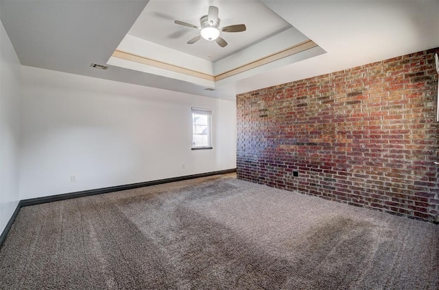 carpeted empty room featuring a tray ceiling, ceiling fan, and brick wall